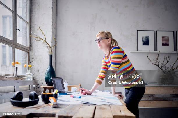portrait of smiling woman standing at desk in a loft looking through window - creative desk stock-fotos und bilder