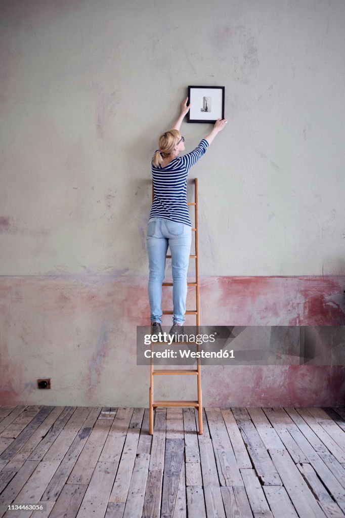 Back view of woman on ladder hanging up picture frame in an unrenovated room of a loft