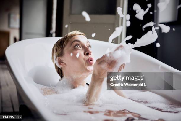 blond woman taking bubble bath blowing foam in the air - bathtub stock-fotos und bilder