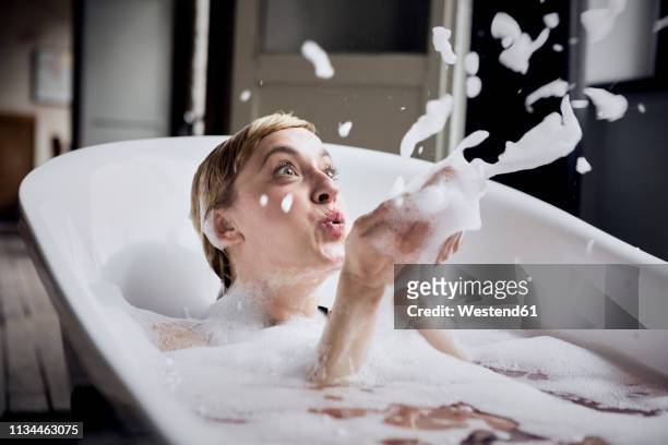 blond woman taking bubble bath blowing foam in the air - bubbelbad stockfoto's en -beelden