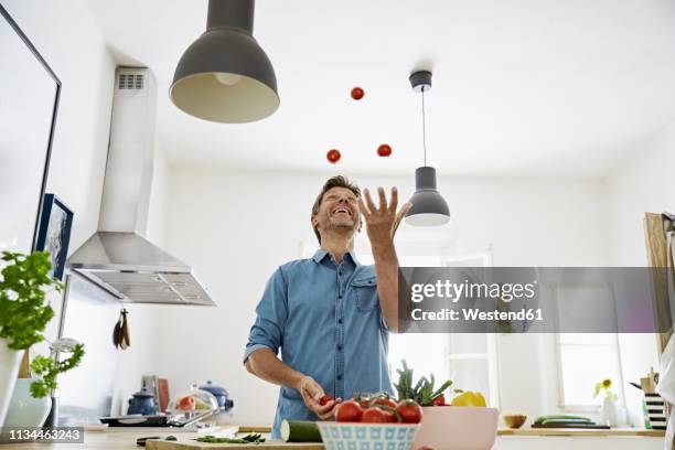 mature man standing in kitchen, juggling with tomatoes - juggling stock pictures, royalty-free photos & images