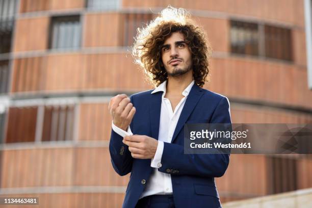 portrait of young fashionable businessman with curly hair wearing blue suit buttoning cuff link - manschettenknöpfe stock-fotos und bilder