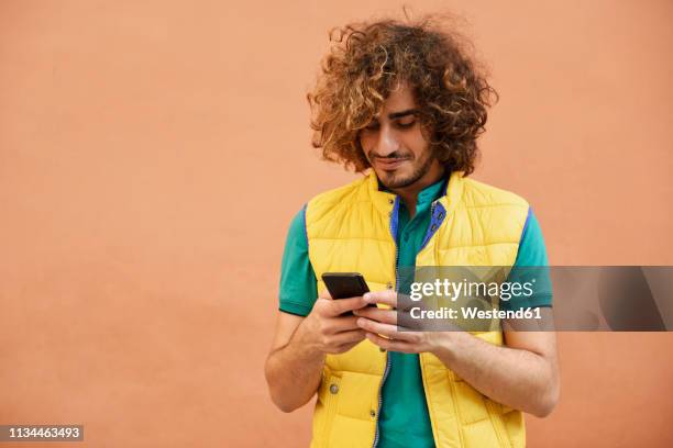 smiling young man with curly hair wearing yellow waistcoat looking at cell phone - chico movil fotografías e imágenes de stock