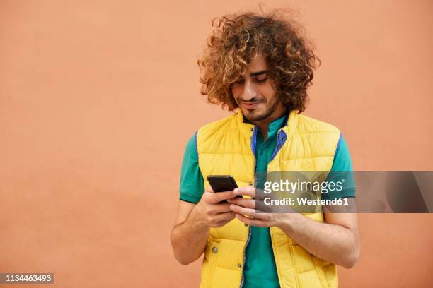 smiling young man with curly hair wearing yellow waistcoat looking at cell phone - phone coloured background stock pictures, royalty-free photos & images