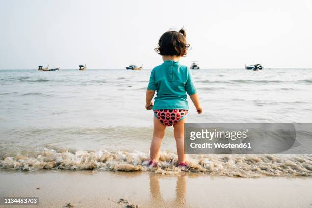 thailand, koh lanta, back view of baby girl wearing uv protection shirt standing at seashore - wasserrand stock-fotos und bilder