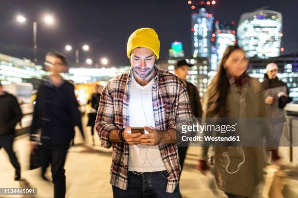 uk, london, smiling man looking at his phone by night with blurred people passing nearby - persona in secondo piano foto e immagini stock