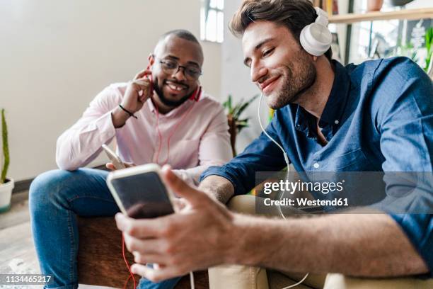 smiling young man showing cell phone to colleague sitting in a loft - 2 men chatting casual office stock-fotos und bilder