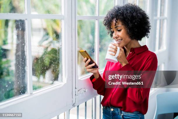 smiling woman with cup of coffee and cell phone standing at the window at home - red blouse fotografías e imágenes de stock