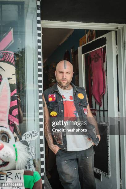 portrait of male punk shop keeper leaning against vintage toy shop doorway - vest stock-fotos und bilder