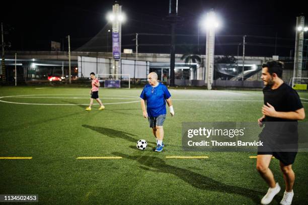 mature man dribbling soccer ball during evening game with friends - flutlicht stock-fotos und bilder