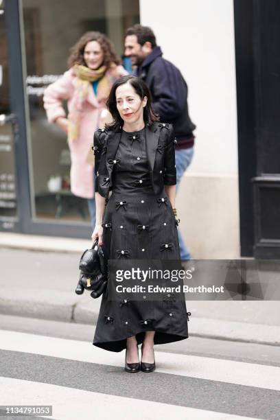 Guest wears lustrous black watered skirt and bolero jacket with bejewelled small bows, a black dog shaped handbag, black pumps , outside Thom Browne,...
