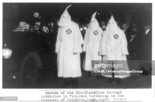 Three members of the Ku Klux Klan in masks and gowns taking part in a night parade. To their left is a car carrying further masked members of the...