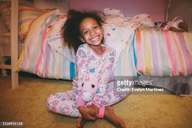 Afrolatina girl sitting on yellow carpet holding a stuff unicorn, smiling at the camera. Colorful bed and pink wall in the background of bedroom.