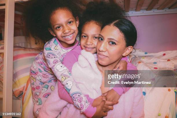 Afrolatina Twin Daughters and Mom embracing, all looking at the camera and wearing pajamas/sleepwear.
