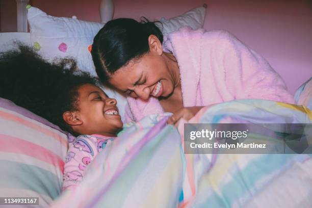 Afrolatina Mom and Daughter laughing candidly, looking away from camera. Mom is wearing a pink robe. Daughter is wearing pajamas.