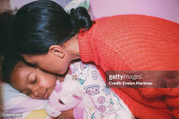 Afrolatina Mom kisses Afrolatina daughter on the cheek. Daughter is wearing pajamas, has her eyes closed, is laying on a bed hugging a stuffed unicorn.