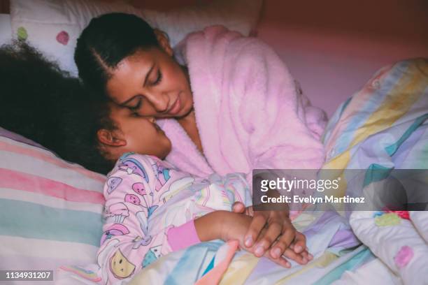 Afrolatina Mom and Daughter with eyes closed, tucked into bed comforter, out of focus. Hands and pajamas in focus.