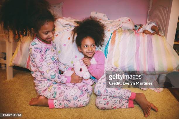 Afrolatina Identical Twins sitting on a yellow carpet, looking away from the camera. Both twins are in pajamas. One twin is looking up and smiling.