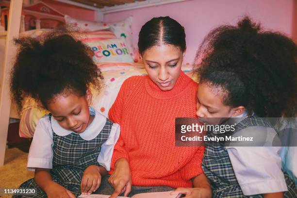 Afrolatina Mom and Twin Daughters looking down at book. Mom and daughters looking away from the camera. Twins are wearing school uniform. Dream in Color pillow and dollhouse in the background of the photo.