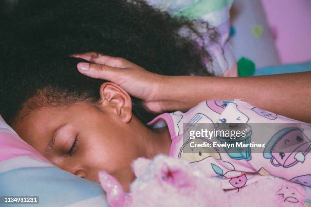Afrolatina girl in pajamas is holding a stuffed unicorn with her eyes closed. Mother's hand is visible in this close-up.