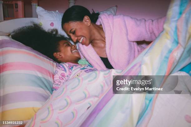 Afrolatina Mom and Afrolatina daughter laughing, tucked into bed comforter. Daughter is wearing pajamas. Mother is wearing a pink robe.