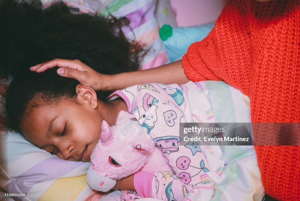 Afrolatina girl in pajamas holding stuffed unicorn with her eyes closed. Mother's face and neck not visible. Mother's hand on daughters head.