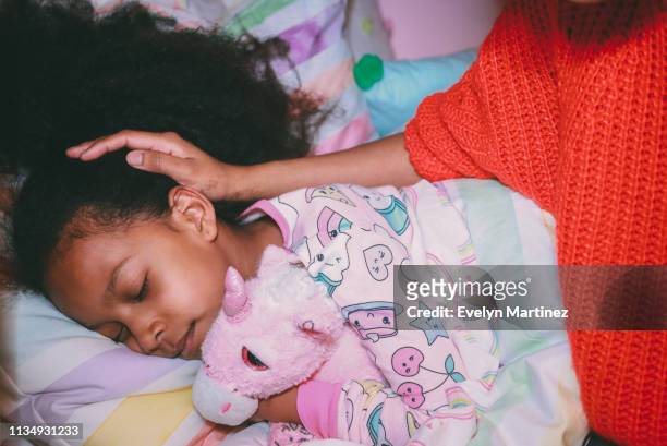 Afrolatina girl in pajamas holding stuffed unicorn with her eyes closed. Mother's face and neck not visible. Mother's hand on daughters head.