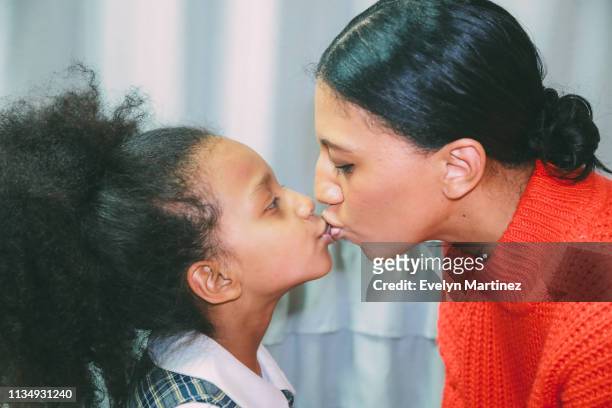 Afrolatina mom kissing Afrolatina daughter on the lips. Mom is wearing hair in a bun. Daughter has her afro in a ponytail and is wearing school uniform.