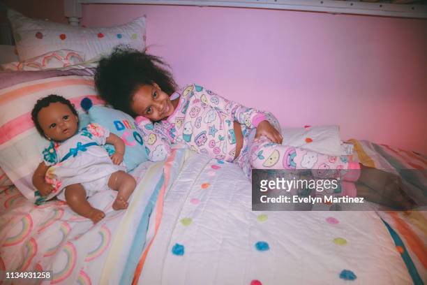 Afrolatina girl in her pajamas on a bed. Pillows and baby doll in the frame. Pink wall in the background.