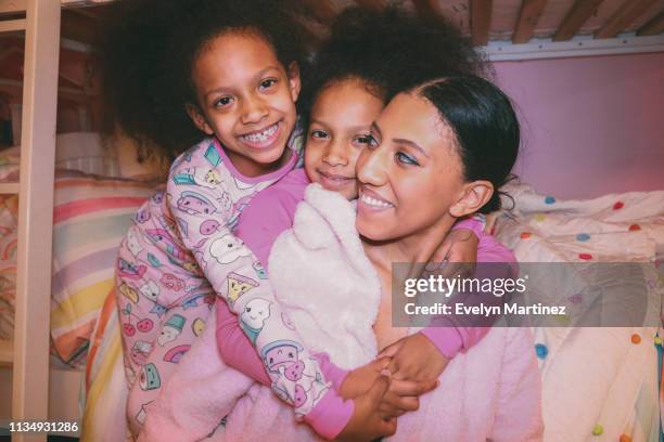 Afrolatina Twin Daughters hugging Afrolatina Mom, everyone is smiling. Daughters looking at camera. Mom is looking away from camera, at her daughters.