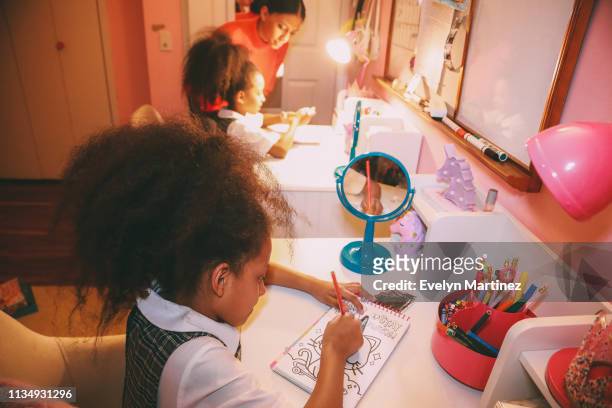 Afrolatina twins coloring at their desks. Mom and other twin out of focus in the background. Closet and bedroom door visible.