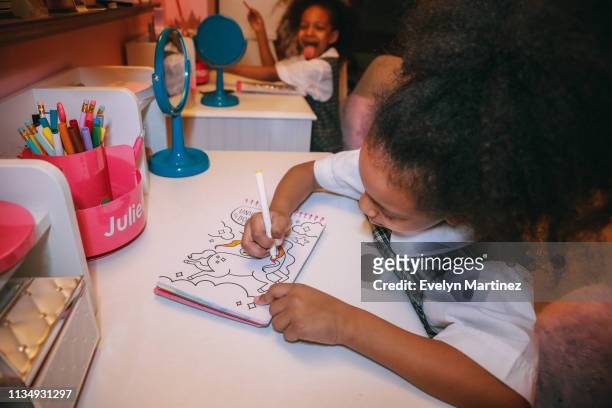 Afrolatina twin girl coloring at her desk. Twin in the background sticking out tongue and smiling. Pencils and desk accessories in the frame.