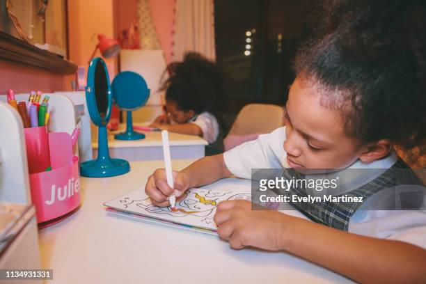 Afrolatina twin sisters sitting at desk coloring. Blue mirrors and pink accessories in the frame. Bedroom window in the background.
