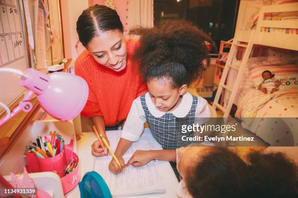 Afrolatina mom and twin daughters doing homework at home. Daughters are in their school uniform. Background is a pink bedroom.
