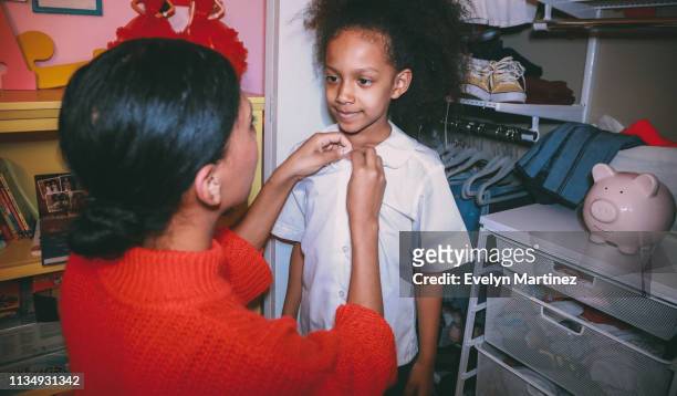 Afrolatina Mom with Afrolatina daughter, by bedroom closet. Mom and daughter looking at each other. Mom has her hands on the buttons of daughter's white shirt.
