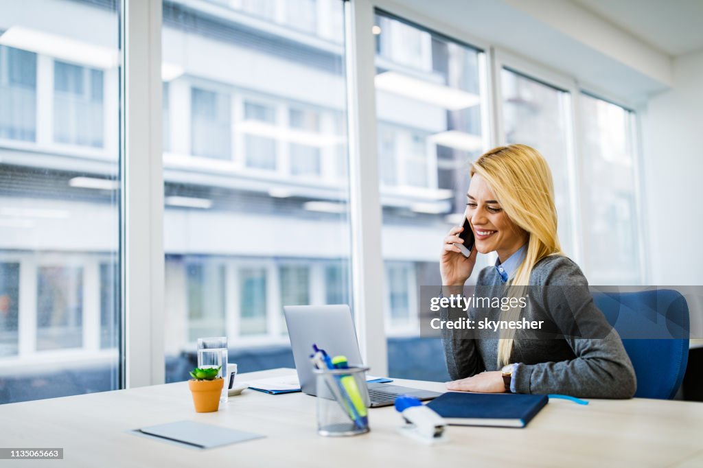 Mulher de negócios feliz que fala no telefone de pilha ao trabalhar no portátil no escritório.