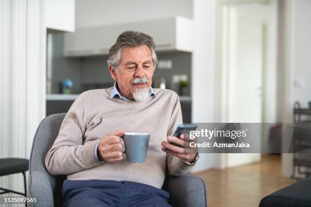 hombre mayor usando el teléfono inteligente y bebiendo café en la sala de estar - un solo hombre mayor fotografías e imágenes de stock