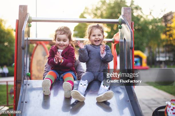 cheerful little girls sitting on a slide at the playground - playground stock pictures, royalty-free photos & images