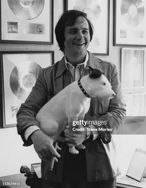 Jerry Weintraub, U.S. Film producer, holding a model of a dog while smoking a cigar, circa 1970. The smiling Weintraub, who stands against a wall...