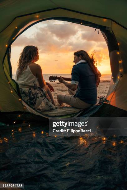 junges erwachsenes paar spielt gitarre bei sonnenuntergang in einem zelt am strand - serenading stock-fotos und bilder