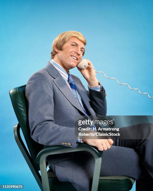 1970s BLOND BUSINESS MAN SITTING IN OFFICE CHAIR TALKING ON TELEPHONE LOOKING AT CAMERA