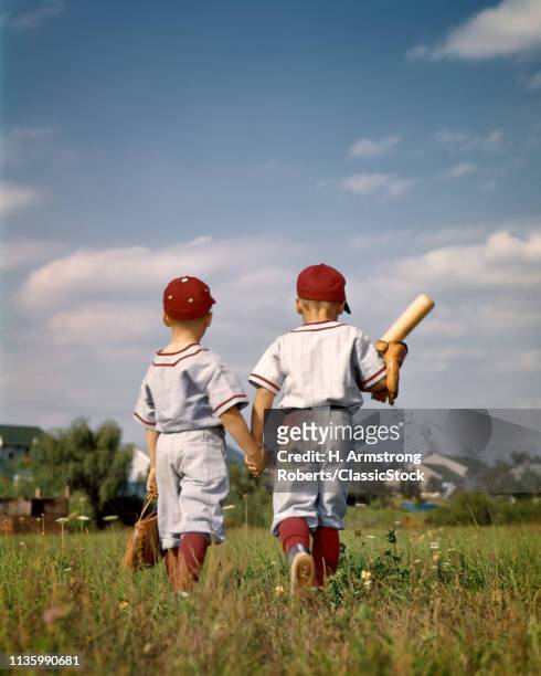 1960s TWO BOYS BROTHERS WEARING LITTLE LEAGUE BASEBALL UNIFORMS HOLDING HANDS WALKING AWAY FROM CAMERA
