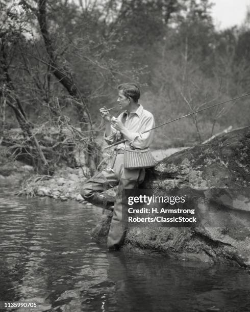 1920s FLY FISHERMAN WEARING WADERS LIGHTING PIPE TAKING A BREAK LEANING ON BOULDER ON BANK OF STREAM