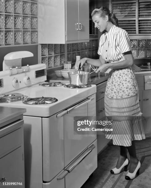 1950s SMILING BLOND HOUSEWIFE WEARING DRESS APRON TWO TONE HIGH HEEL SHOES COOKING ON MID-CENTURY MODERN ELECTRIC KITCHEN RANGE