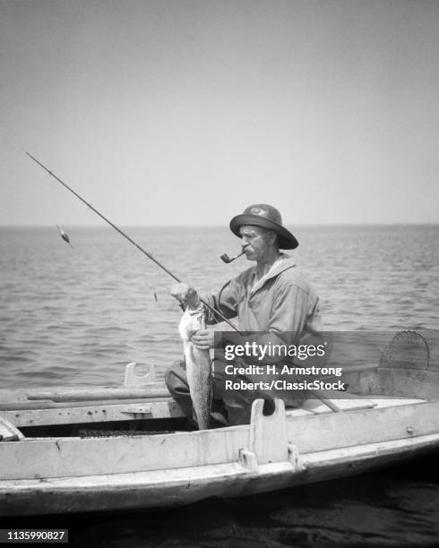 1920s MAN BARNEGAT BAY COMMERCIAL FISHERMAN SITTING IN DUCK BOAT SMOKING PIPE REMOVING FISHHOOK FROM FISH CATCH NEW JERSEY USA