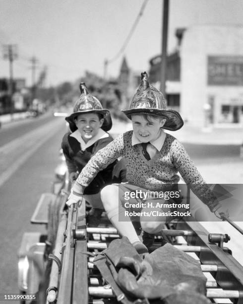 1930s TWO SMILING EXCITED PRETEEN BOYS WEARING FIREMAN SAFETY HELMETS CLIMBING SITTING RIDING PLAYING ON BACK OF A FIRE TRUCK