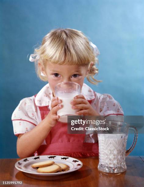 1950s PRETTY LITTLE CURLY HAIR BLOND GIRL LOOKING AT CAMERA DRINKING MILK FROM GLASS AND PITCHER WITH A PLATE OF COOKIES