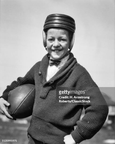 1920s SMILING FRECKLE FACE PRETEEN BOY WEARING EARLY LEATHER HELMET AND SHAWL-COLLAR SWEATER HOLDING FOOTBALL LOOKING AT CAMERA