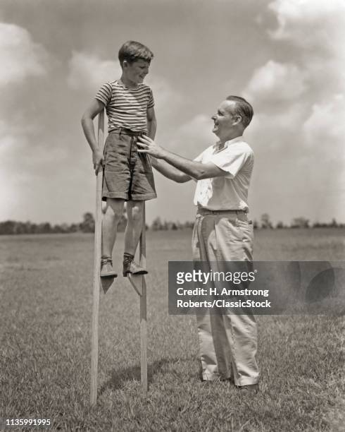 1930s SMILING MAN FATHER PARENT TEACHING ENCOURAGING SUPPORTING SHOWING BOY SON HOW TO BALANCE AND WALK ON TALL WOOD STILTS