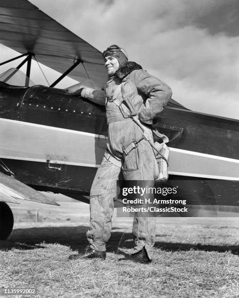 1940s PORTRAIT SMILING MAN AVIATOR PILOT STANDING BY BIPLANE WEARING CANVAS FLYING SUIT HELMET GOGGLES AND PARACHUTE